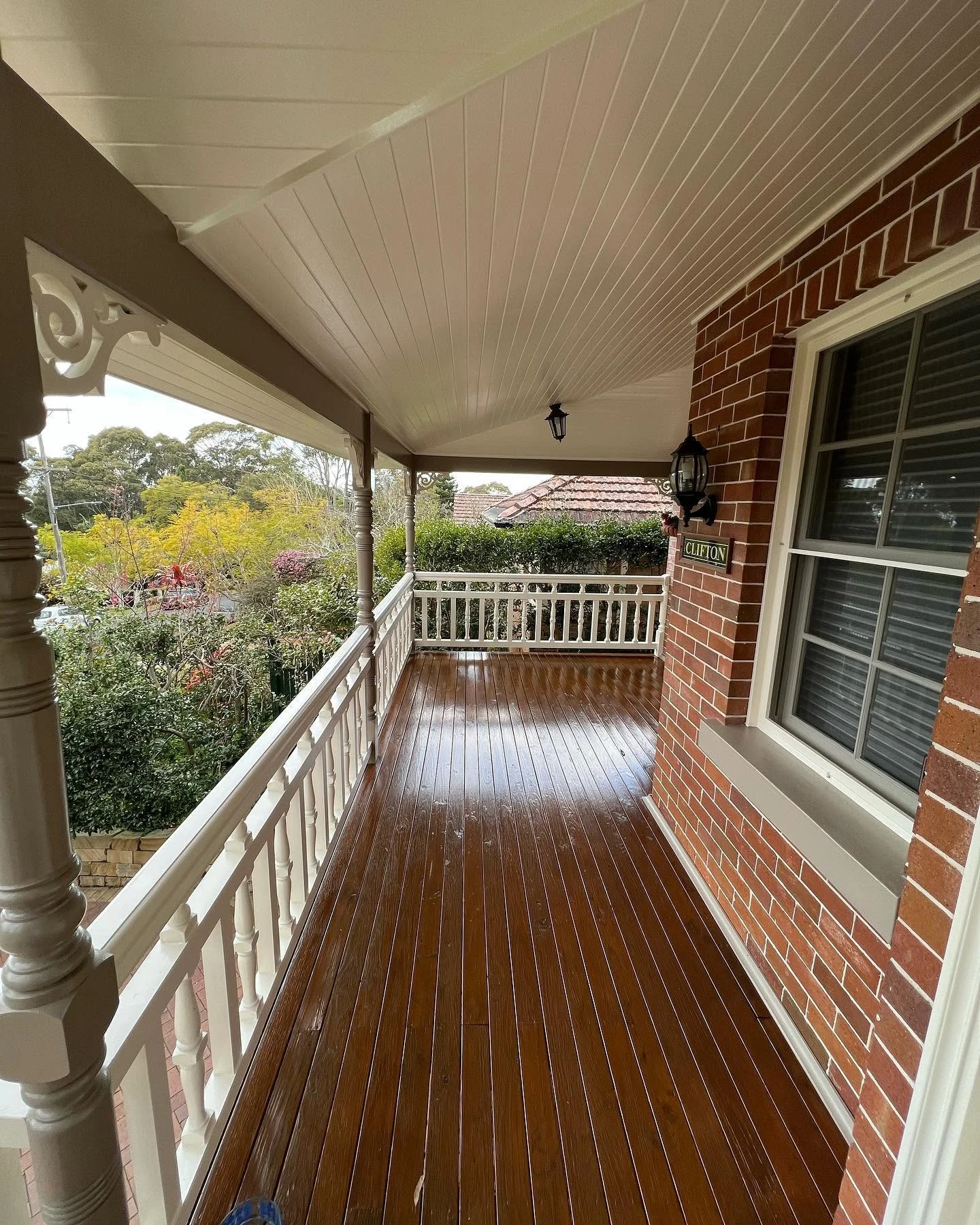Covered outdoor veranda with white ceiling and balustrades beside a brick wall and timber floor.